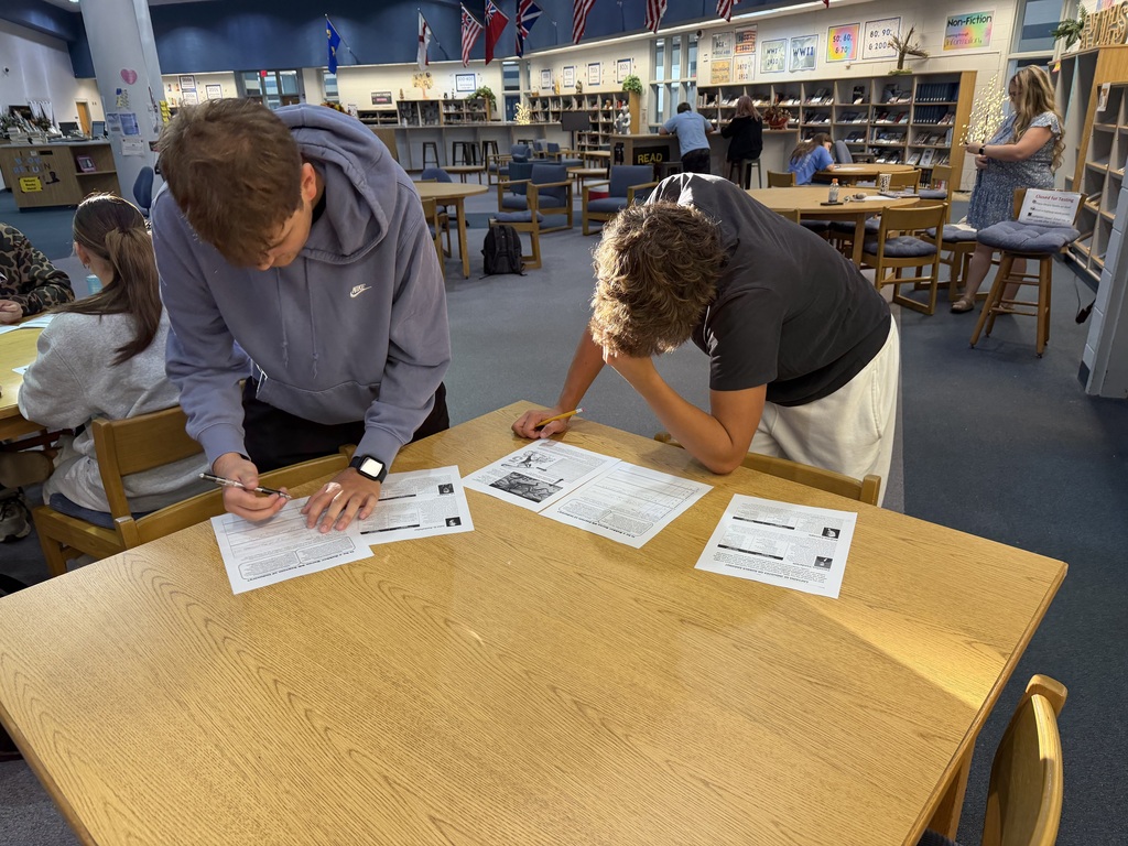 Two people work on papers at a table in a library. Other people sit at tables behind them.