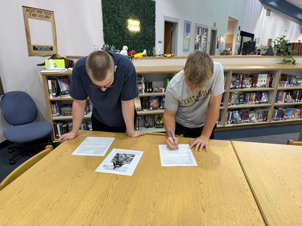 Two people signing papers at a desk in a library. Behind them are shelves with books.
