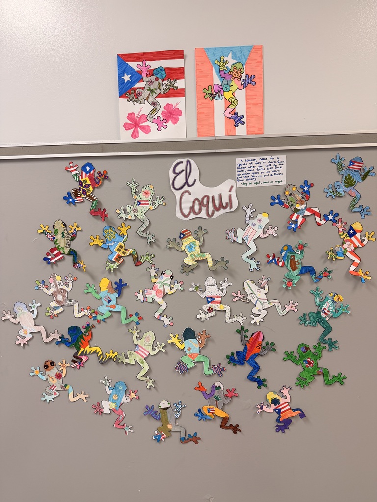 A gray bulletin board displays numerous colorful frog cutouts, forming a circular shape. Above the board, two small flags are attached.