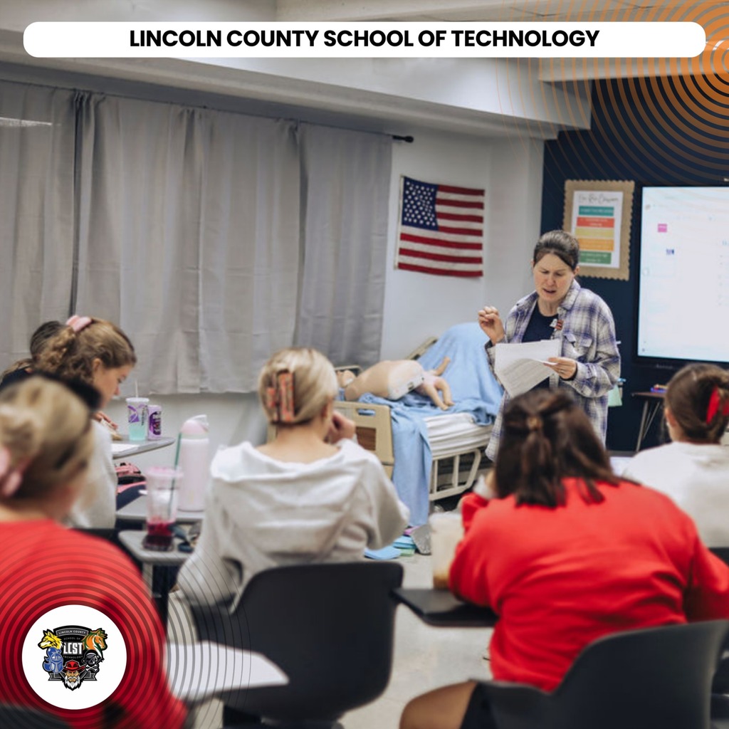 A classroom setting with students seated at tables. A teacher stands at the front, possibly demonstrating a lesson.
