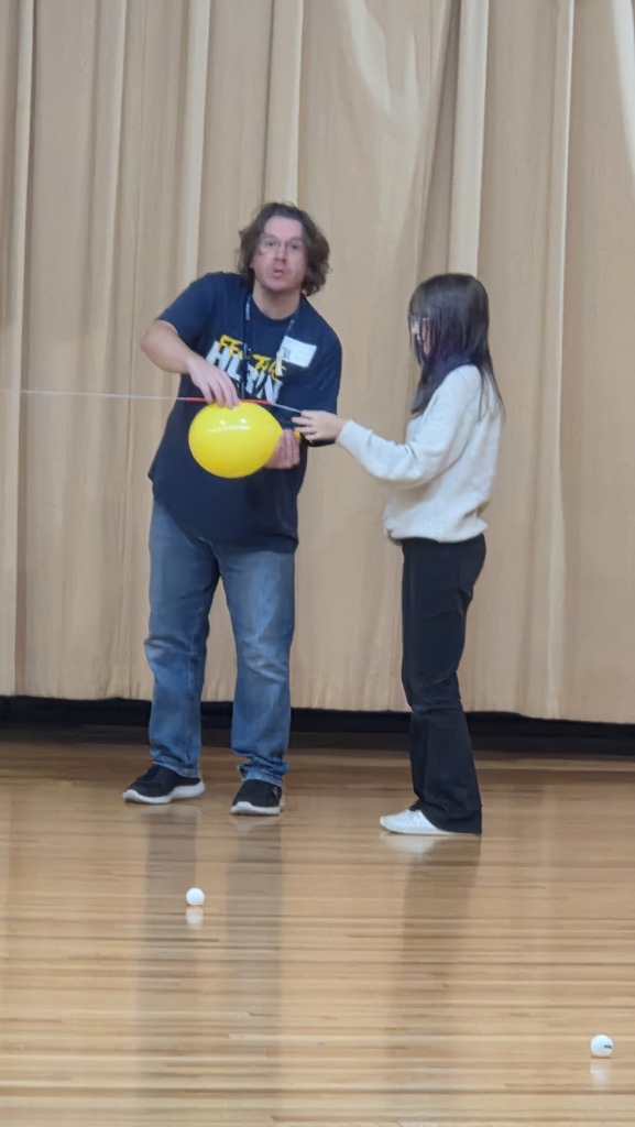 Two people in a room with a curtain. One holds a yellow ball while the other reaches to touch it.