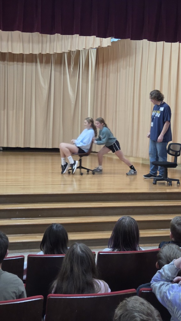 Three people on a stage, two seated and one standing. Audience seated in chairs in front.