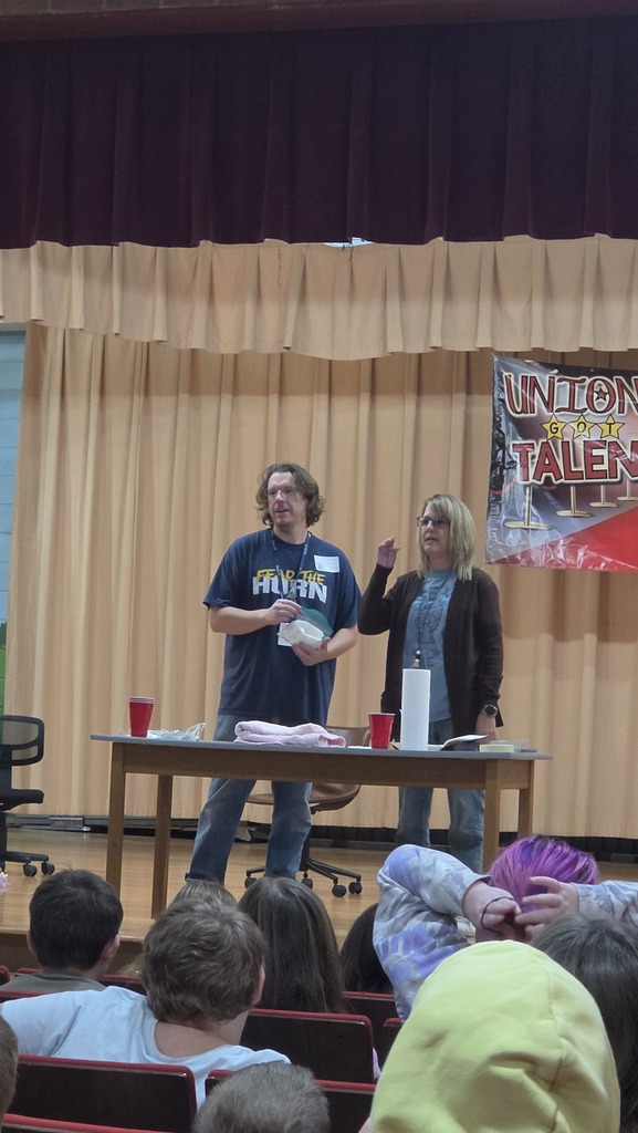 Two people on stage with a banner behind them, one holds a paper, the other a cup. Audience seated in front, looking on.