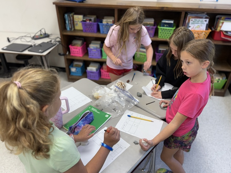 Four young students work together around a gray activity table inside a classroom setting. In the foreground, a girl with blonde hair in a ponytail and a light green shirt writes on a worksheet beside a student in a bright pink t-shirt holding a piece of chalk or rock. Across the table, a girl with long blonde hair in a light pink shirt leans over to inspect scientific specimens, near another student writing with a pencil. Shelves filled with colorful storage bins and books line the back wall.