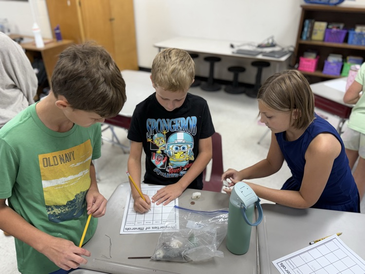 Three students work together at a classroom desk examining mineral specimens during a science activity. A student on the left in a green Old Navy t-shirt and a student in the middle wearing a black SpongeBob t-shirt write observations on a "Properties of Minerals" activity worksheet. A girl on the right in a sleeveless navy blue dress tests a mineral specimen with a tool near a green water bottle. In the background, other desks and classroom furniture are visible.