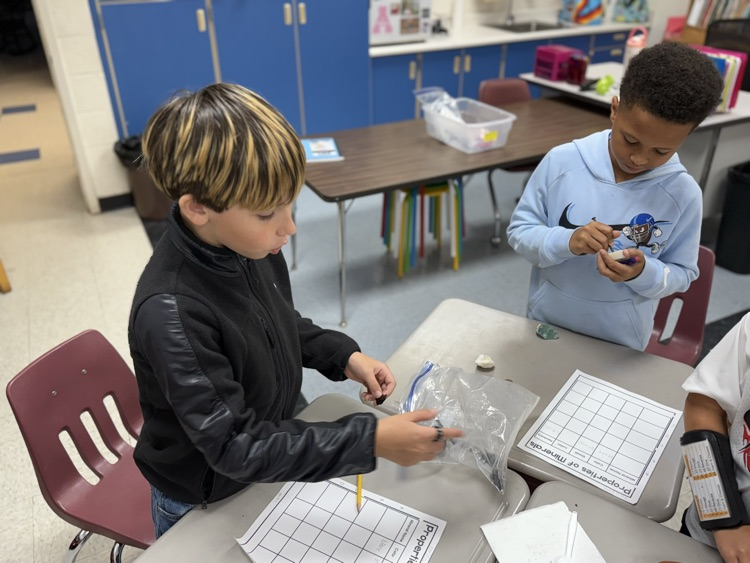 Two male elementary students working at classroom desks on a "Properties of Minerals" science activity. On the left, a student with dark hair and blonde highlights wearing a black jacket reaches into a plastic bag containing mineral specimens while holding a pencil over his activity grid. On the right, a student in a light blue Nike hoodie holding a mineral sample examines it closely. In the background, classroom furniture and storage cabinets are visible.