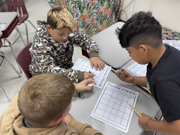 An overhead view of three male elementary students collaborating at a desk on a science activity. Two students on the left, one wearing a camouflage hoodie and another wearing a light brown hoodie, examine rock and mineral specimens while holding them over their desks. On the right, a student wearing a black t-shirt holds a pencil and writes observations on a grid paper worksheet titled "Properties of Minerals".
