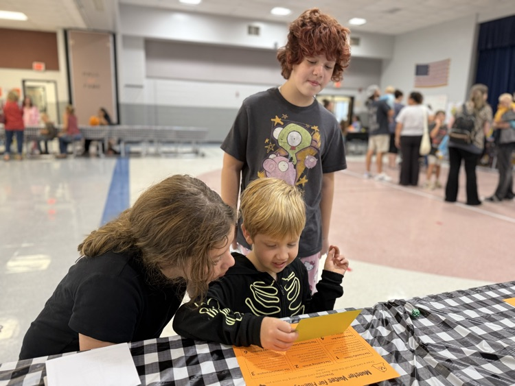 A child with curly reddish hair wearing a gray shirt with cartoon graphics stands behind two children sitting at a table with a black and white checkered tablecloth. The young boy in the center wears a black skeleton zip-up hoodie and reads an orange activity paper titled "Monster Voices for Fluency" with help from a girl with long brown hair leaning in beside him. In the background, other adults and children gather in a spacious school cafeteria or gymnasium with a US flag hanging on the back wall.
