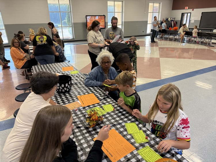 A high-angle view of students and adults gathered around a table with a black and white checkered tablecloth during a school literacy event in a gymnasium or cafeteria. In the foreground, two students and an adult sit at the table looking at orange and yellow instructional activity sheets titled "Monster Voices for Fluency". In the center, an adult woman with glasses and short gray hair sits with students at the table while an adult man in a gray shirt stands nearby. In the background, other parents and students walk through the room past windows and display screens.