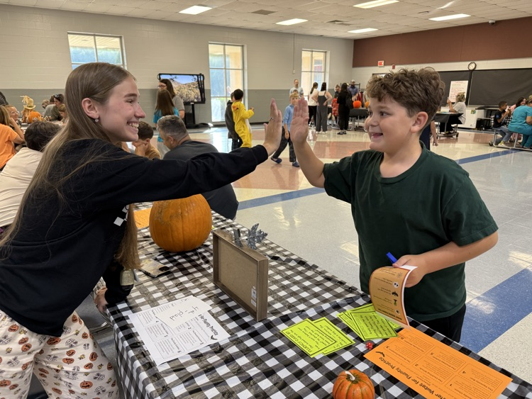 A young girl with long brown hair leaning across a table with a black and white checkered tablecloth to high-five a young boy wearing a green t-shirt during a school event. On the table sits a large pumpkin, a small pumpkin, and activity sheets titled "Monster Voices for Fluency Practice". In the background, other adults and children gather in a spacious school cafeteria or gymnasium.