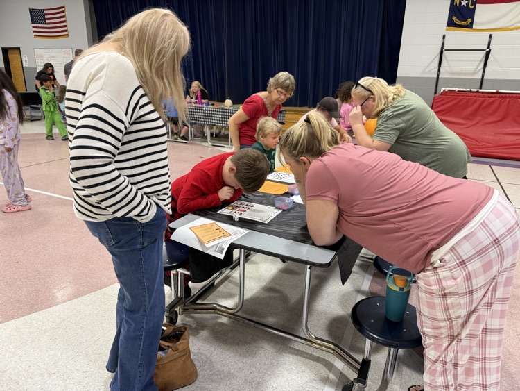 A side-angle view of adults and students gathered around small cafeteria tables during an educational activity in a school gymnasium or cafeteria. On the left, a woman with long blonde hair wearing a black-and-white striped long-sleeve shirt and blue jeans leans forward to look at activity papers on a table. Across from her, another woman wearing a pink shirt and pink plaid pajama pants leans over the table near a young boy in a red shirt who is focused on a worksheet. In the background, other adults and children interact around tables, with a dark blue stage curtain and a North Carolina state flag visible on the wall.