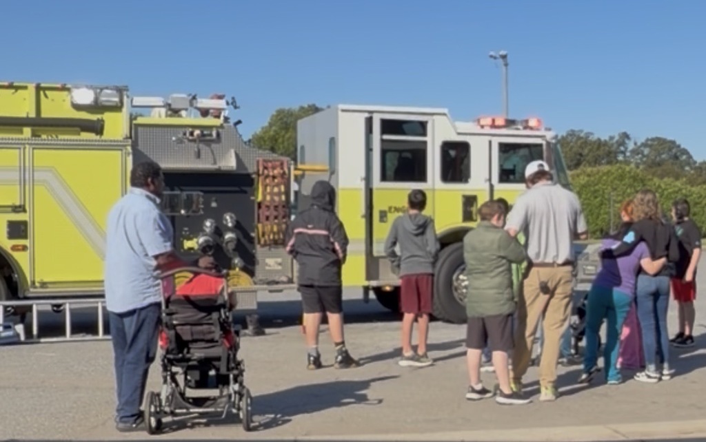 Several people gather in front of a yellow fire truck with a man pushing a stroller.