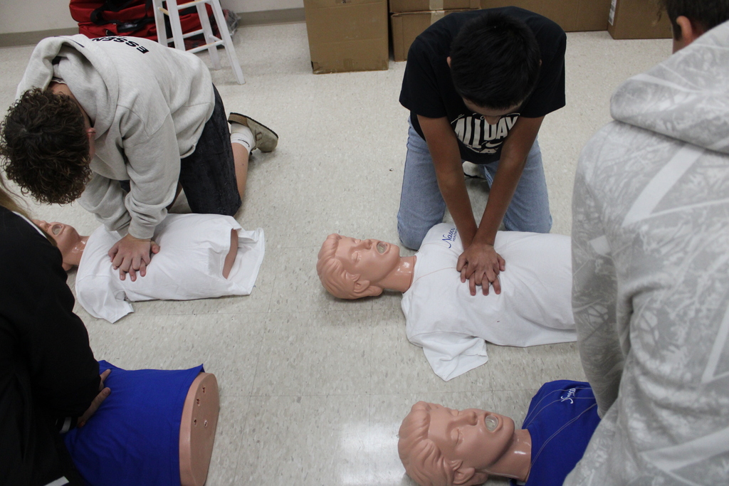 Several people in hoodies and shorts practice CPR on mannequins in a room with white tile flooring.