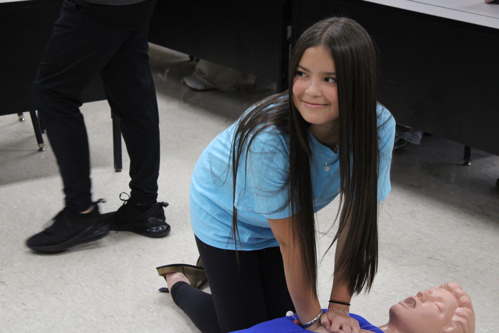 A girl kneels, practicing CPR on a mannequin, wearing a blue shirt and black pants. A person stands behind her in black pants and shoes.