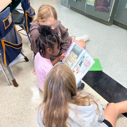 Three girls sit on floor, one reads from book to others, in a classroom.