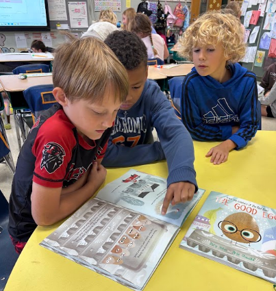 Three young children look at books at a yellow table, focusing on the pages in front of them.