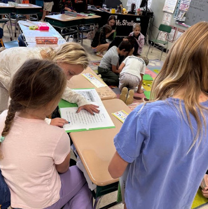 Children in a classroom setting. One child reading aloud to two other children.
