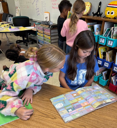 Two girls examine an open book on a table in a classroom. Nearby, other children are seated and standing.