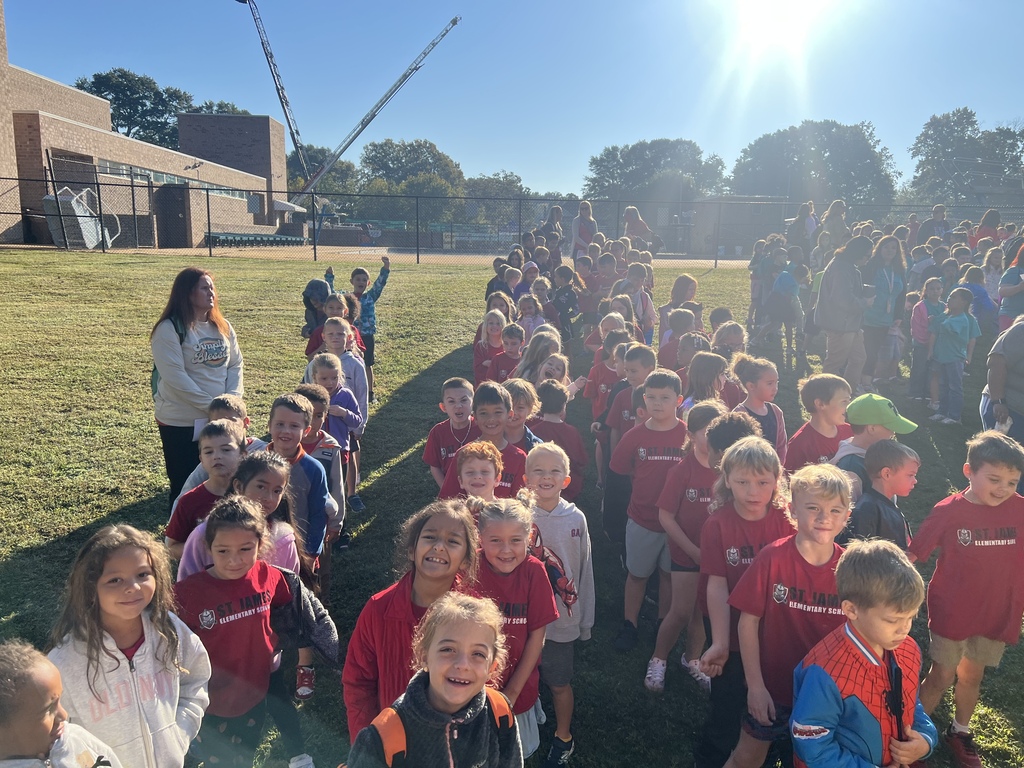 A group of children in red shirts stand in a line on a grassy field with trees.