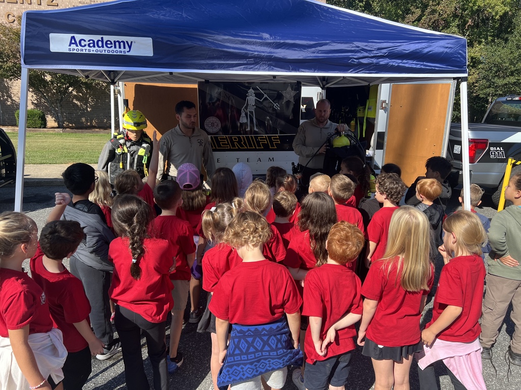 A group of children in red shirts and hats are gathered around a canopy tent.