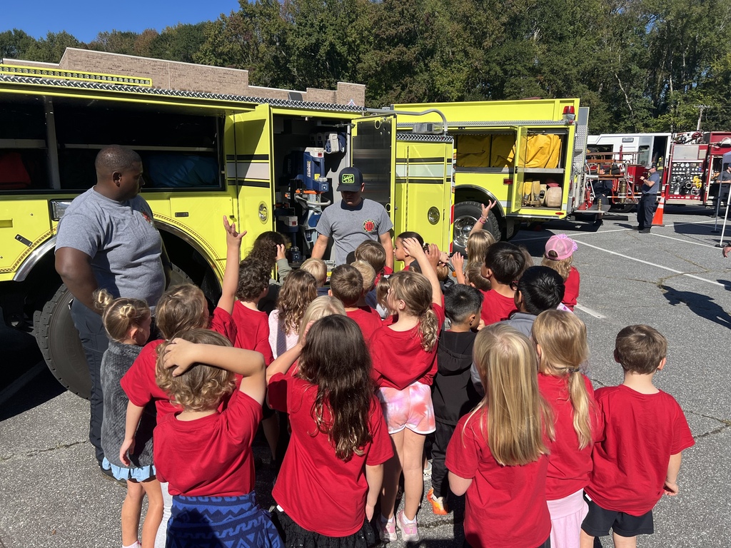 A group of children in red shirts gathers around a yellow fire truck. A man in a gray shirt stands inside the open door.