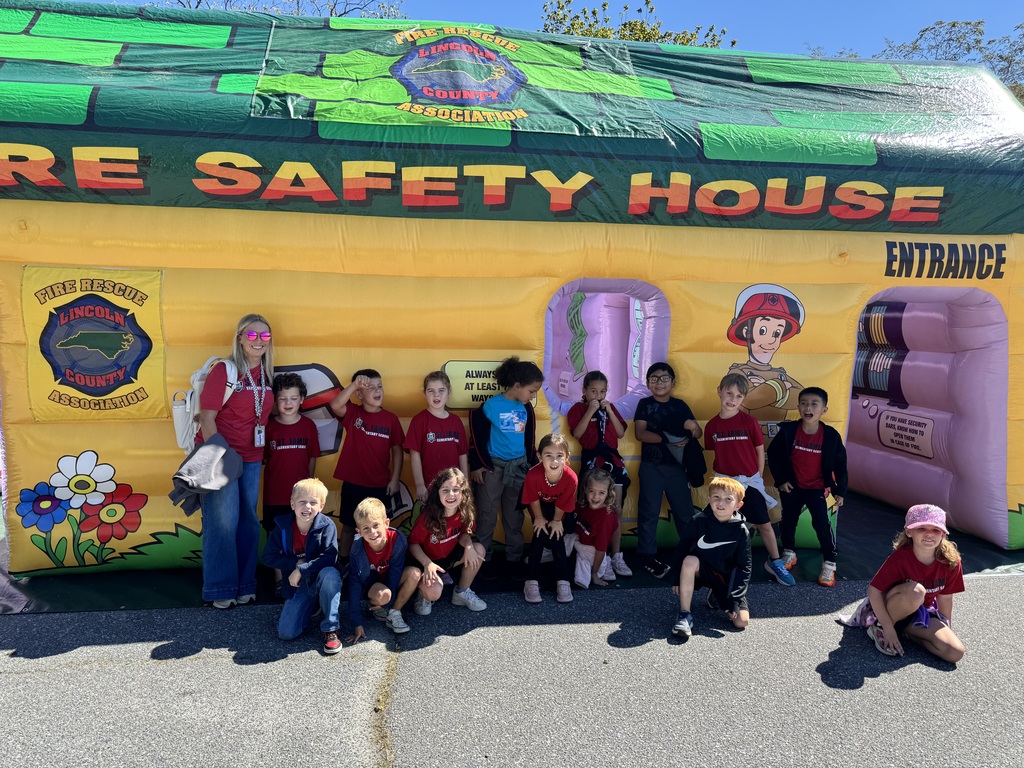 A group of children and two adults standing in front of a large inflatable structure with text reading "Fire Safety House".