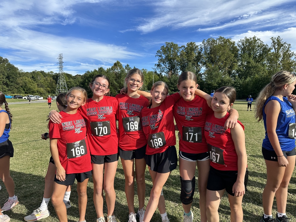 Seven girls wearing matching red shirts with numbers stand together on a grassy field, with trees in the background.