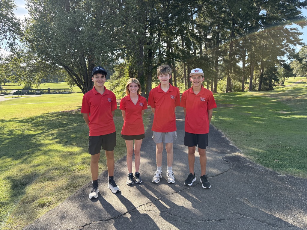 Four individuals in red shirts, black shorts, and sneakers standing together on a path at a golf course.