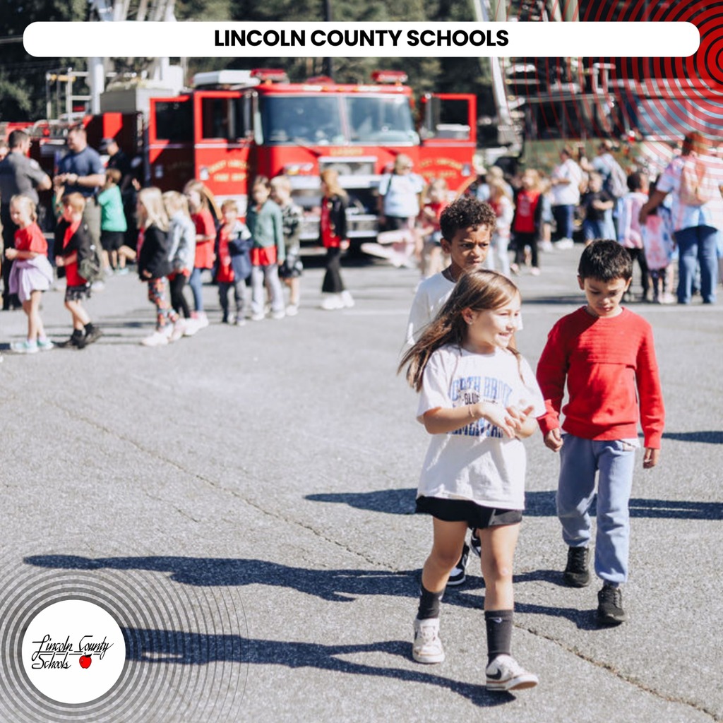Children walking in a line in front of a fire truck. A school logo is in the lower left corner.