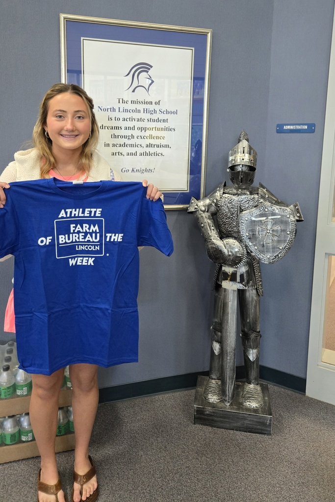 A woman holds a blue t-shirt in front of a knight statue in a room with blue walls and a large framed quote.