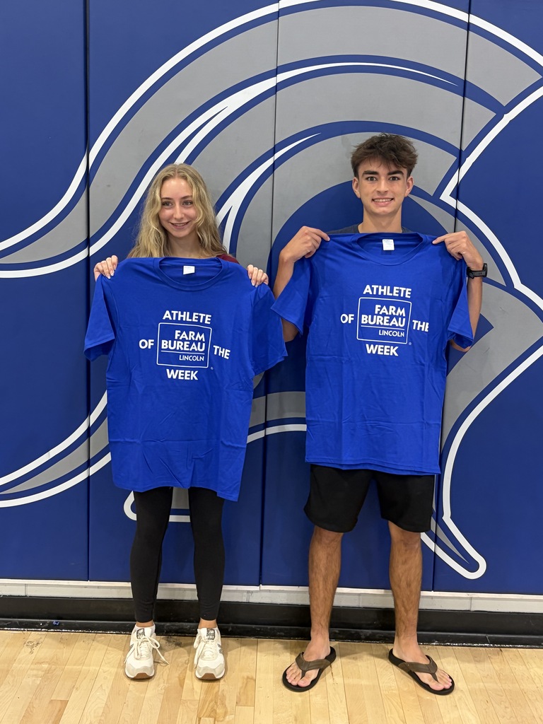 Two individuals stand against a blue and white backdrop, holding blue shirts with white text.