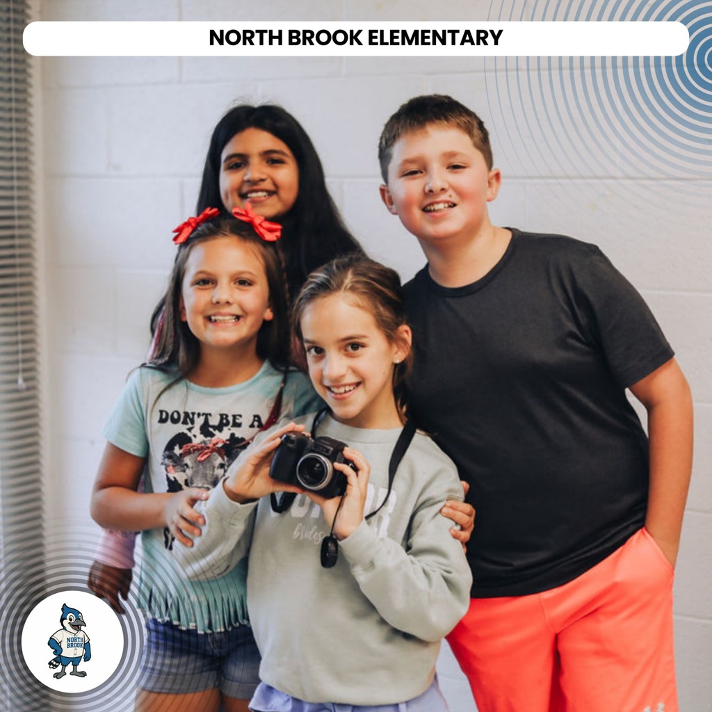 Four children stand together, one holding a camera. Two girls wear light-colored tops, and one has a red bow. A boy wears orange pants and a black shirt.