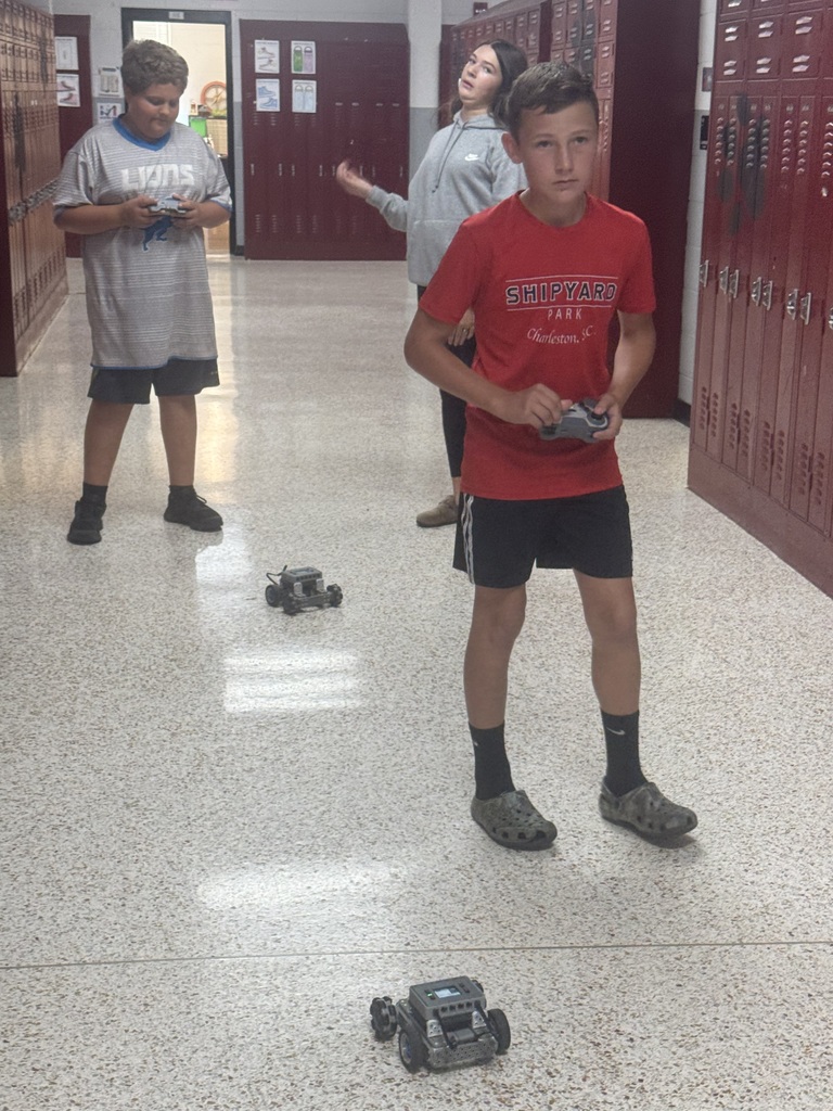 Three children in a school hallway, one holding a remote control and the others holding remotes.