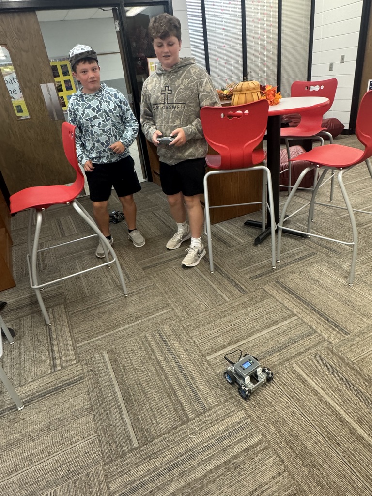 Two individuals in a room with chairs, a table, and a robot on the floor.