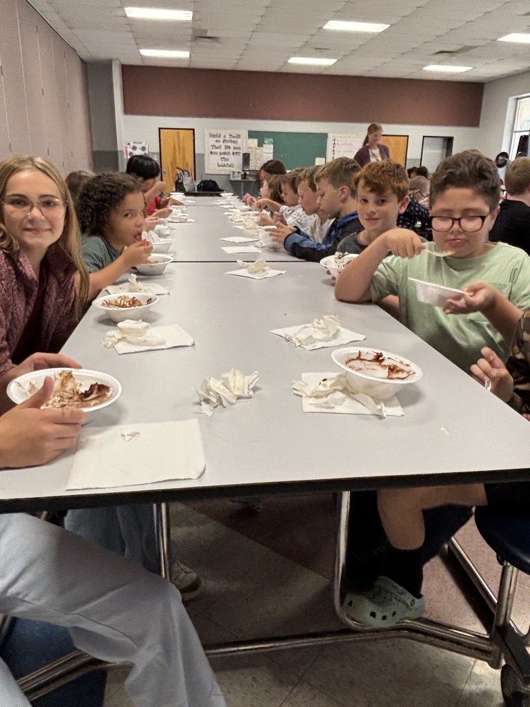 Elementary school students seated along a long cafeteria table eating ice cream sundaes out of bowls. One student wearing glasses and a green shirt eats with a spoon in the foreground, while adult staff stand in the background near classroom doors and display boards.