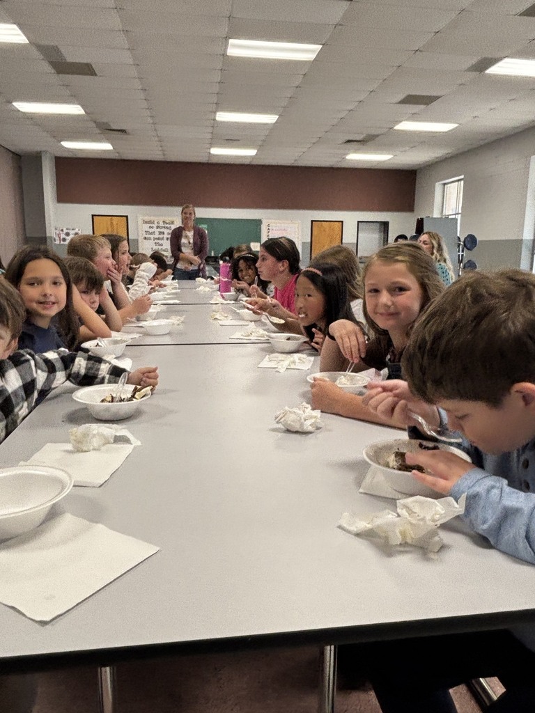 A classroom cafeteria setting where elementary school students sit along a long gray table, eating dessert from bowls using plastic spoons. Adult staff members stand in the background near a chalkboard and classroom door.
