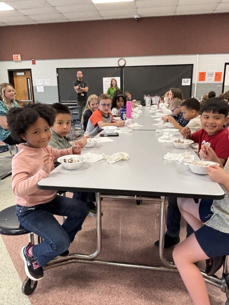 A young student sitting at a cafeteria table smiles while holding a spoon over a bowl of ice cream. Other elementary students sit along both sides of the long gray table eating sundaes, while adult staff stand in the background near chalkboards and classroom doors.