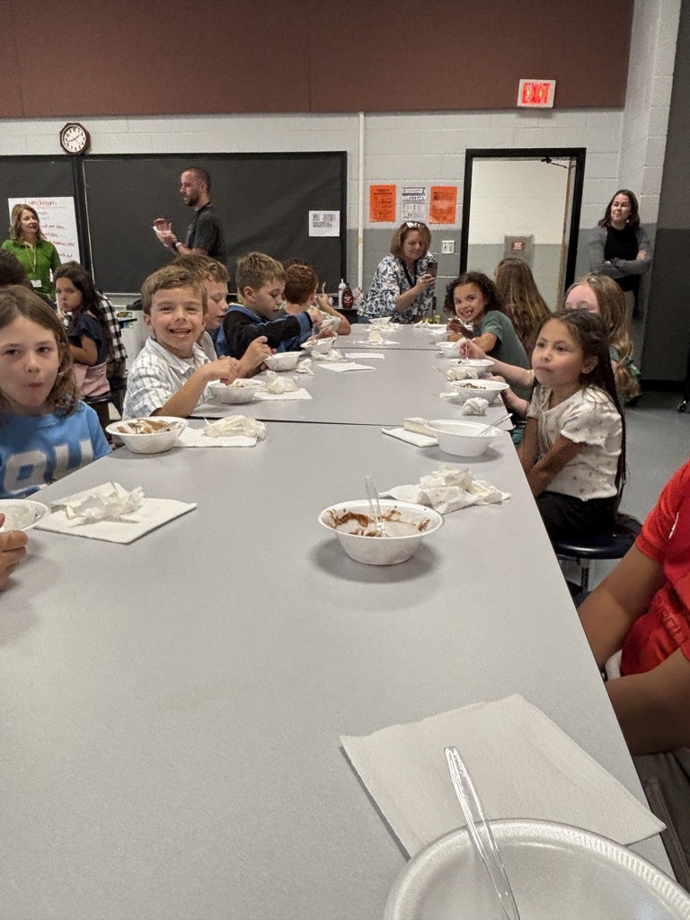 Elementary school students sit at a long gray cafeteria table smiling and eating ice cream sundaes out of bowls with plastic spoons. Adults stand in the background of the cafeteria space near whiteboards, an exit door, and posters.