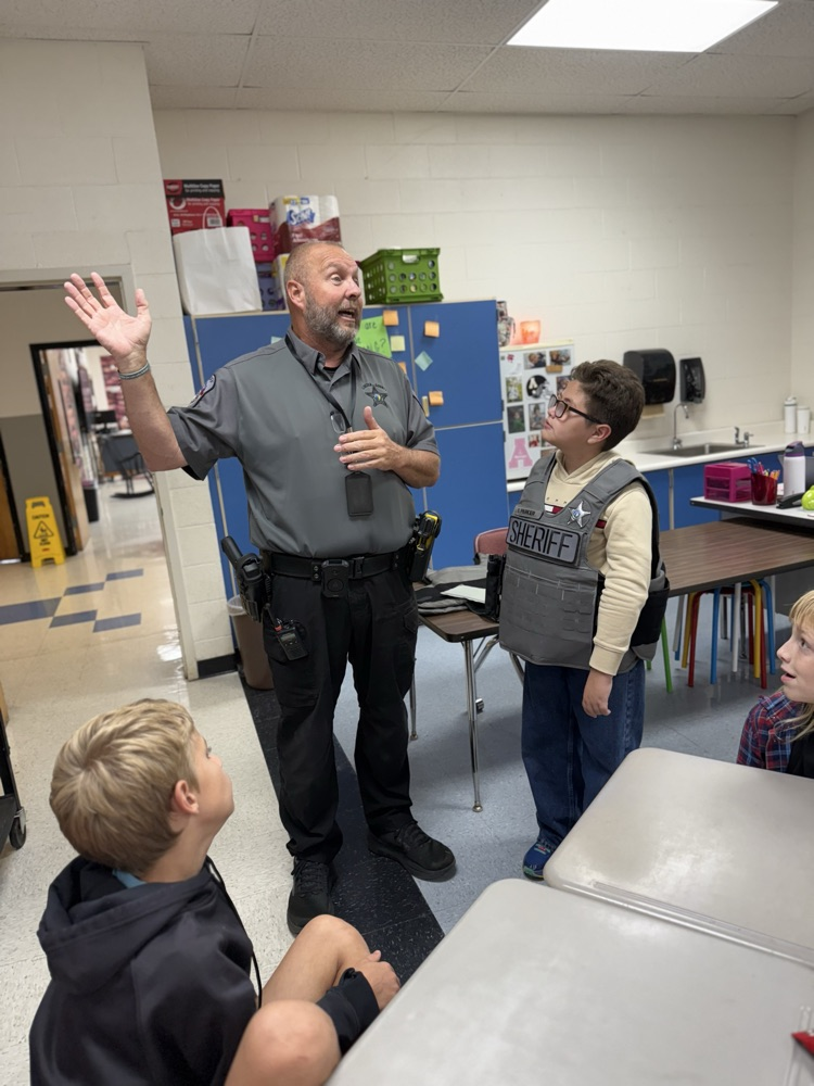 A police officer in a gray short-sleeve uniform shirt and black pants stands in a classroom gesturing with open hands as he speaks to students. To his right, a young boy wearing glasses and a gray tactical vest labeled "SHERIFF" with a star badge looks up at him. Other elementary students sit at desks watching and listening to the officer.