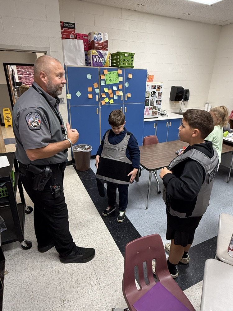 A police officer wearing a gray short-sleeve uniform shirt with a sheriff patch stands on the left, gesturing with his thumb toward two elementary school students standing in a classroom. The boy on the left looks down while wearing a dark gray protective vest. The boy on the right wears a gray tactical vest marked "SHERIFF" as he looks toward the officer.