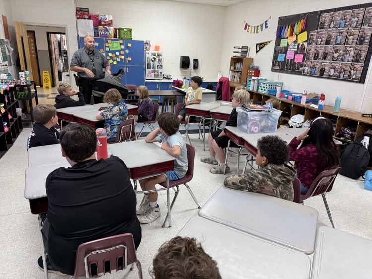 A police officer in a gray short-sleeve uniform shirt stands at the front of a classroom holding a gray tactical vest while presenting to a room of elementary school students sitting at their desks watching him. On the wall in the background, there is a poster reading "BELIEVE" and a bulletin board featuring student photographs.