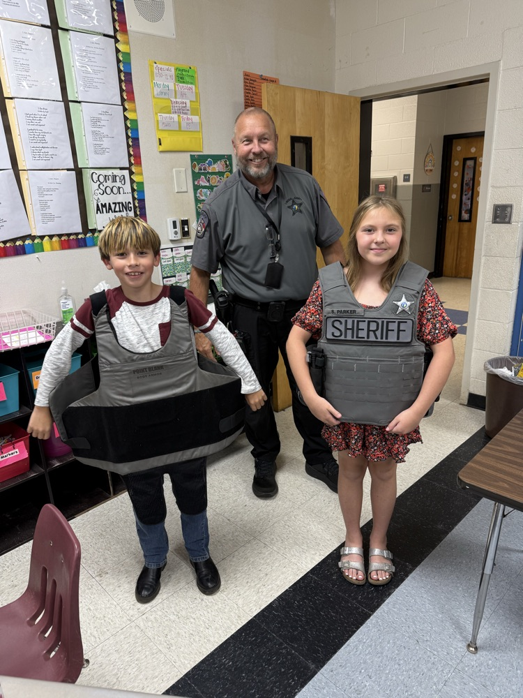 A smiling police officer in a gray short-sleeve uniform shirt stands in a classroom behind two elementary school students who are trying on tactical vests. The boy on the left smiles while holding open a wide, gray protective vest. The girl on the right stands smiling, wearing a gray tactical vest labeled "SHERIFF" with a star badge.