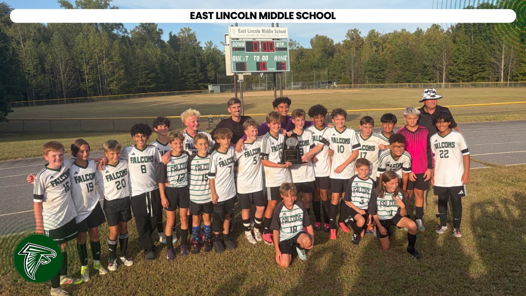 A group of children in matching uniforms poses for a photo on a grassy field with a scoreboard in the background.