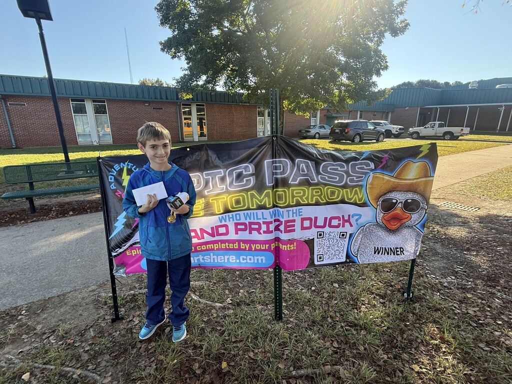 A boy in blue stands beside a banner with a duck image and text, near a school building.