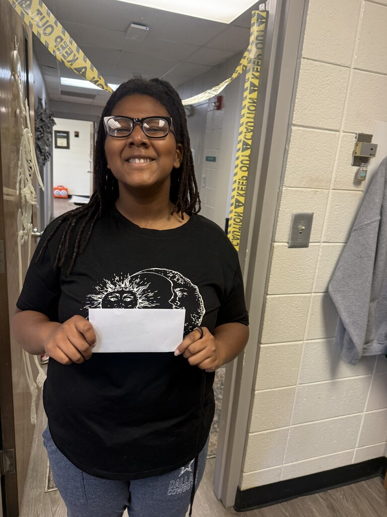 A person holds a paper in both hands, smiling, wearing a black t-shirt and glasses, standing near a door.