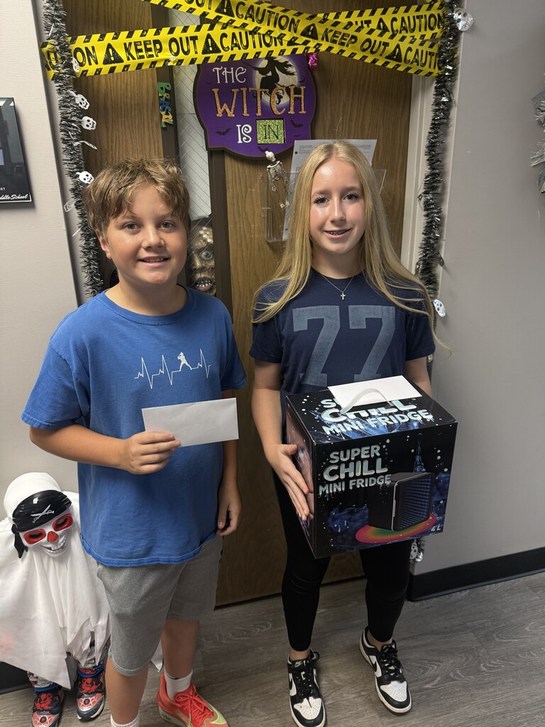 Two children stand in front of a door with Halloween decorations. One holds a box, and the other holds a paper.