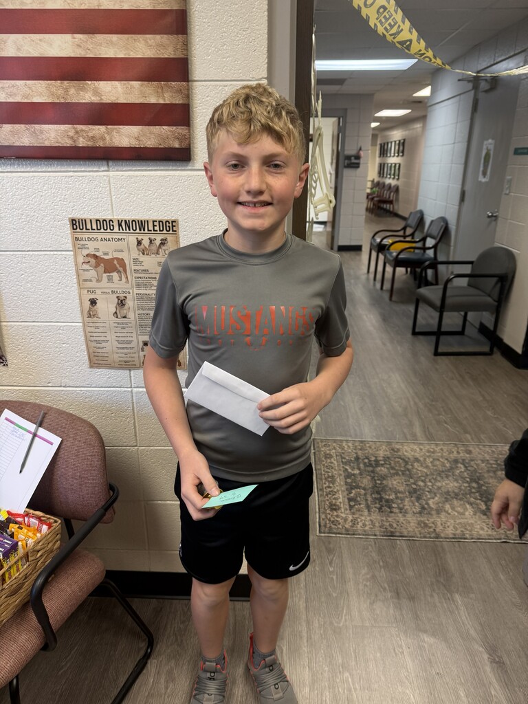 A young boy stands in a hallway, holding a paper and a green card, wearing a gray shirt and black shorts.
