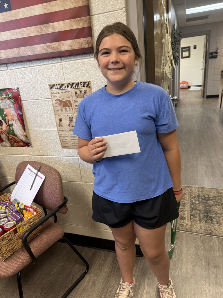 A young girl stands in a hallway, holding a piece of paper and smiling. She wears a blue shirt and black shorts.