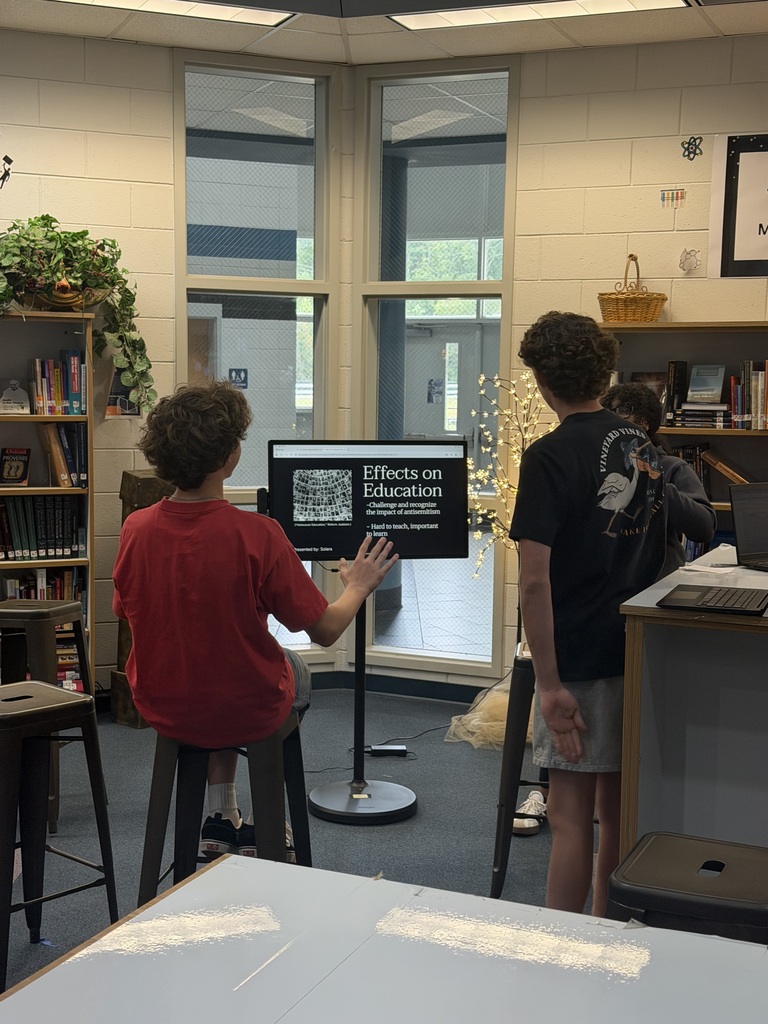 Three students in a library area. Two boys focus on a screen while the third prepares a laptop.