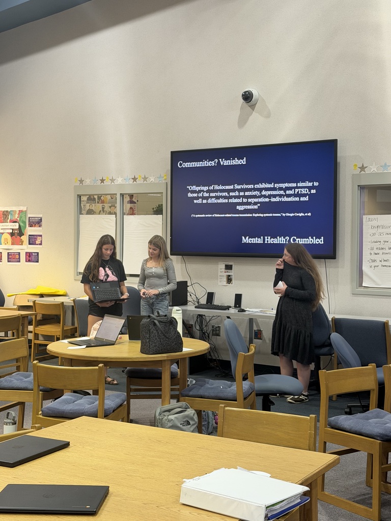 Three women stand near a table with laptops in a classroom. A monitor displays a presentation.